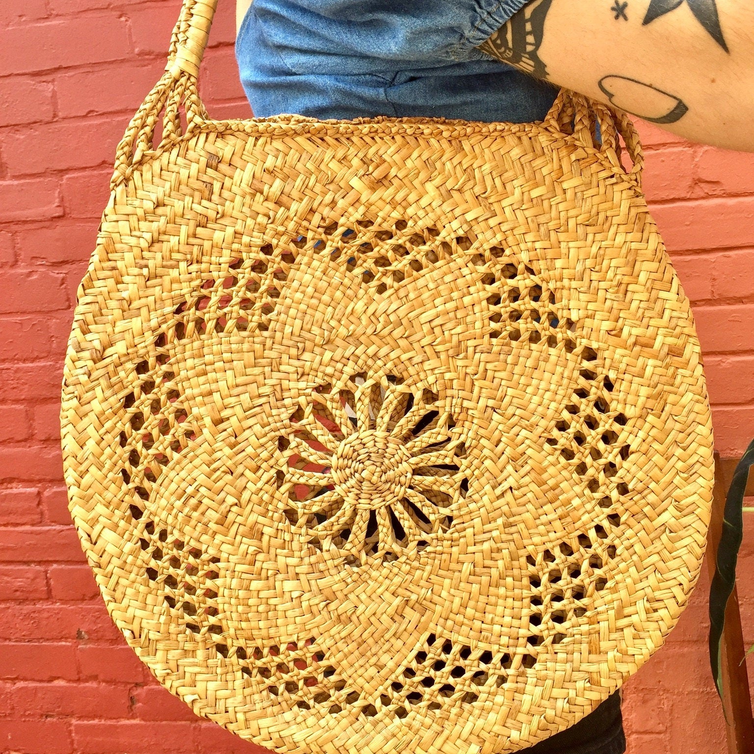 Round woven straw bag with intricate floral pattern design hanging on a person's tattooed arm against a red brick wall background.
