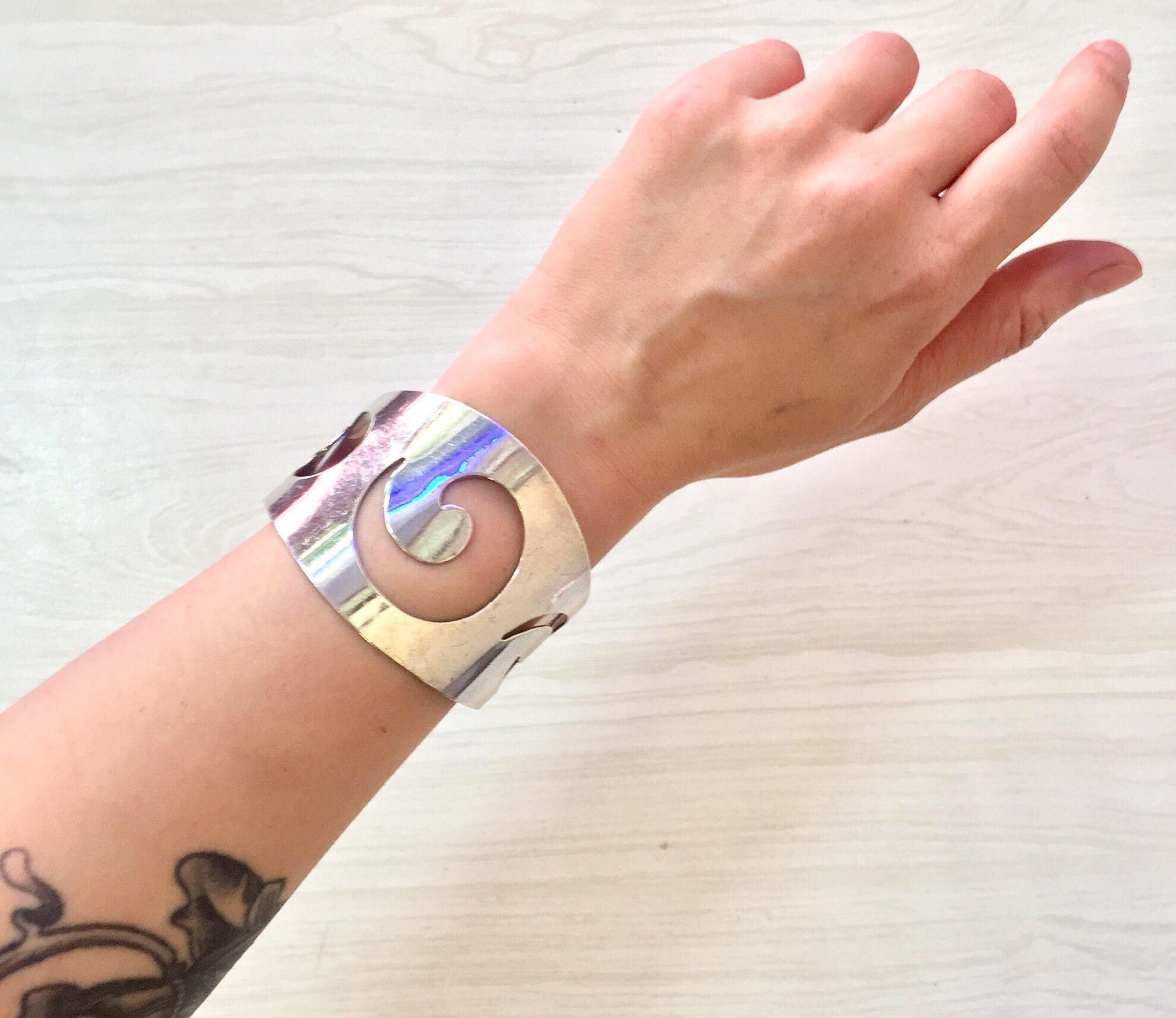 Vintage sterling silver cuff bracelet with iridescent swirl design on a tattooed wrist against a white textured background.