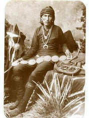 Vintage black and white photograph of a Native American man in traditional clothing sitting outdoors, wearing a concho belt and multiple necklaces while holding pottery vessels.