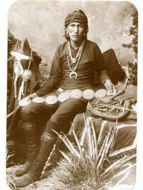 Vintage black and white photograph of a Native American man in traditional clothing sitting outdoors, wearing a concho belt and multiple necklaces while holding pottery vessels.
