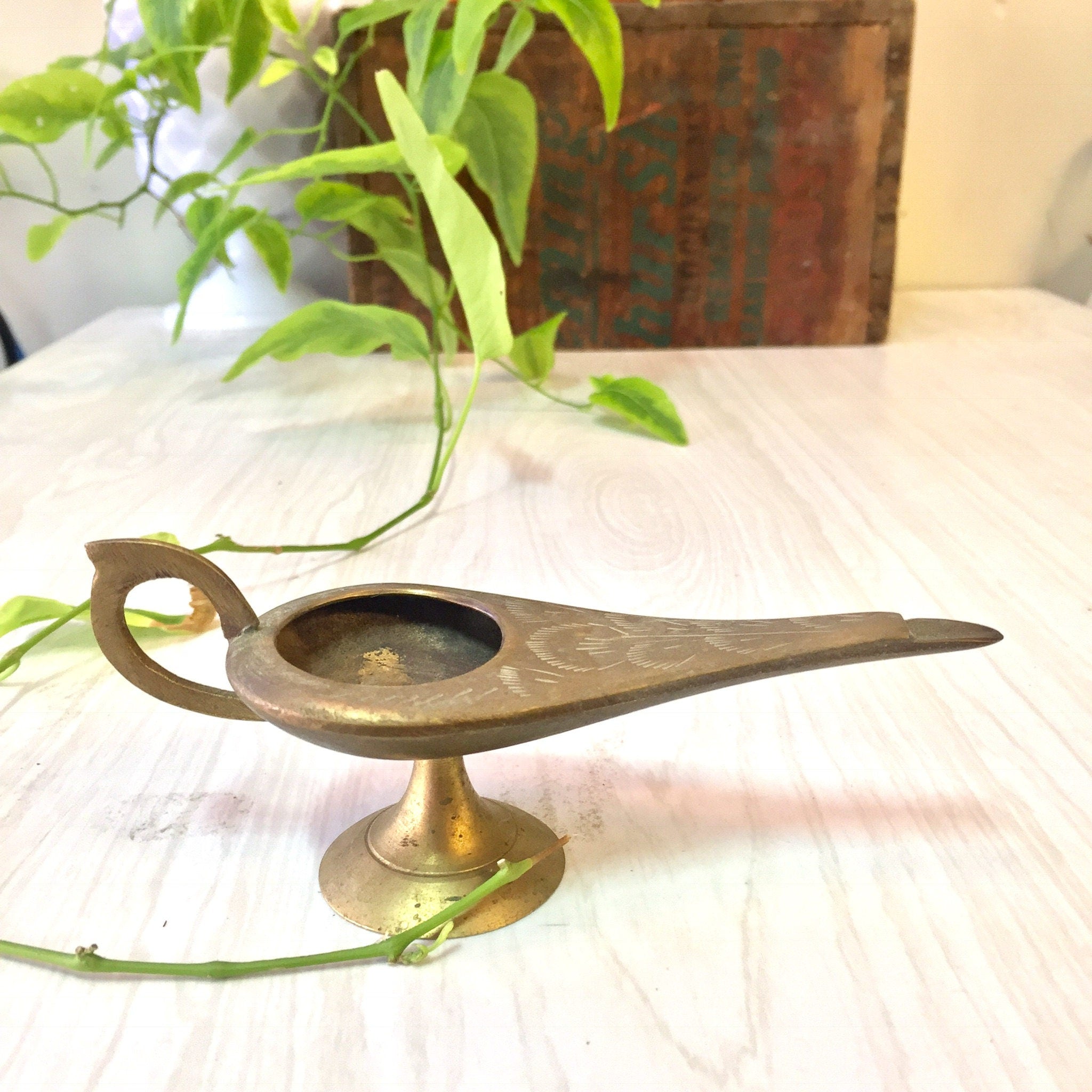 Vintage brass genie lamp shaped incense burner with etched designs sitting on white fabric next to green plant leaves and vintage box in background