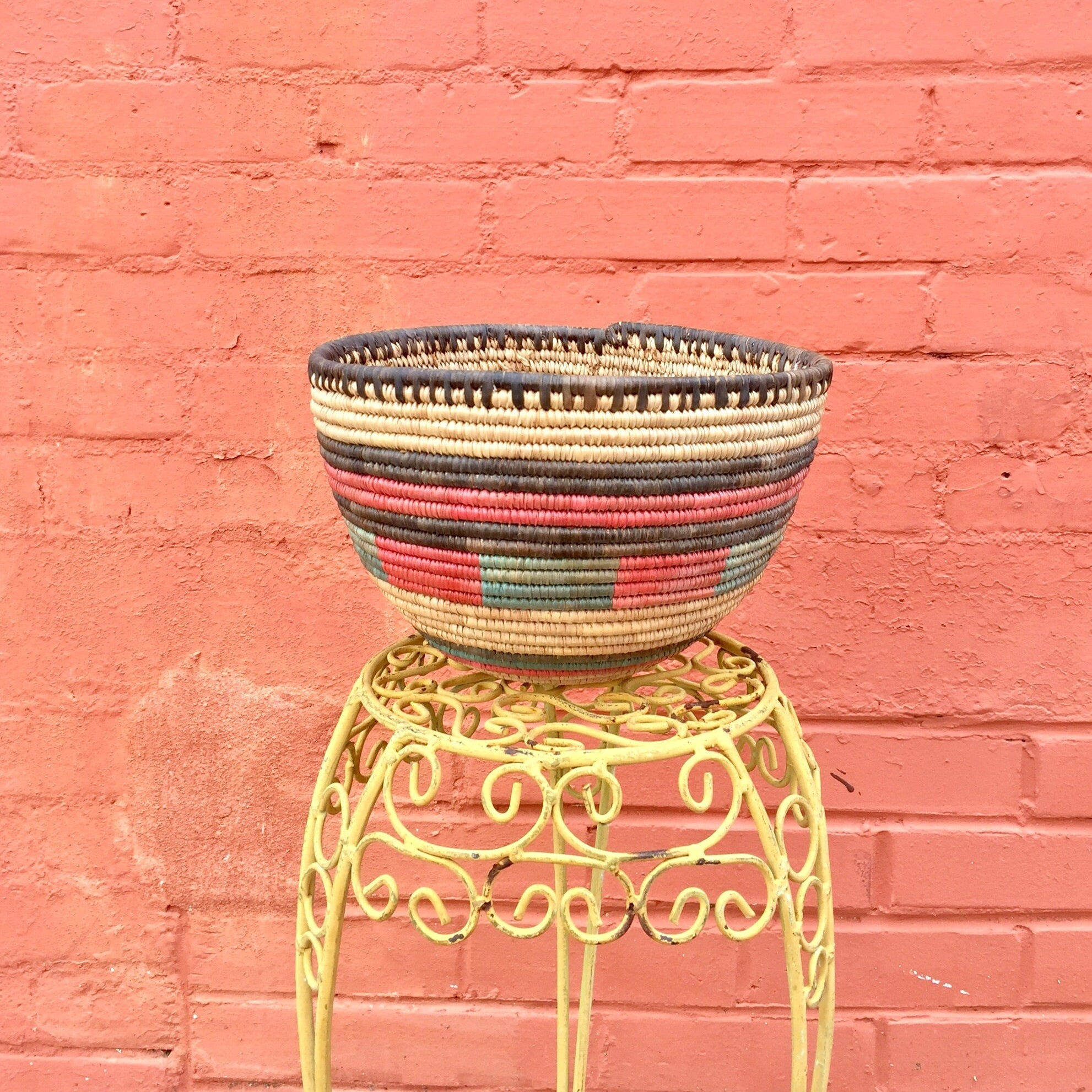 Colorful woven basket on yellow metal stand against pink brick wall, handmade bohemian vintage home decor