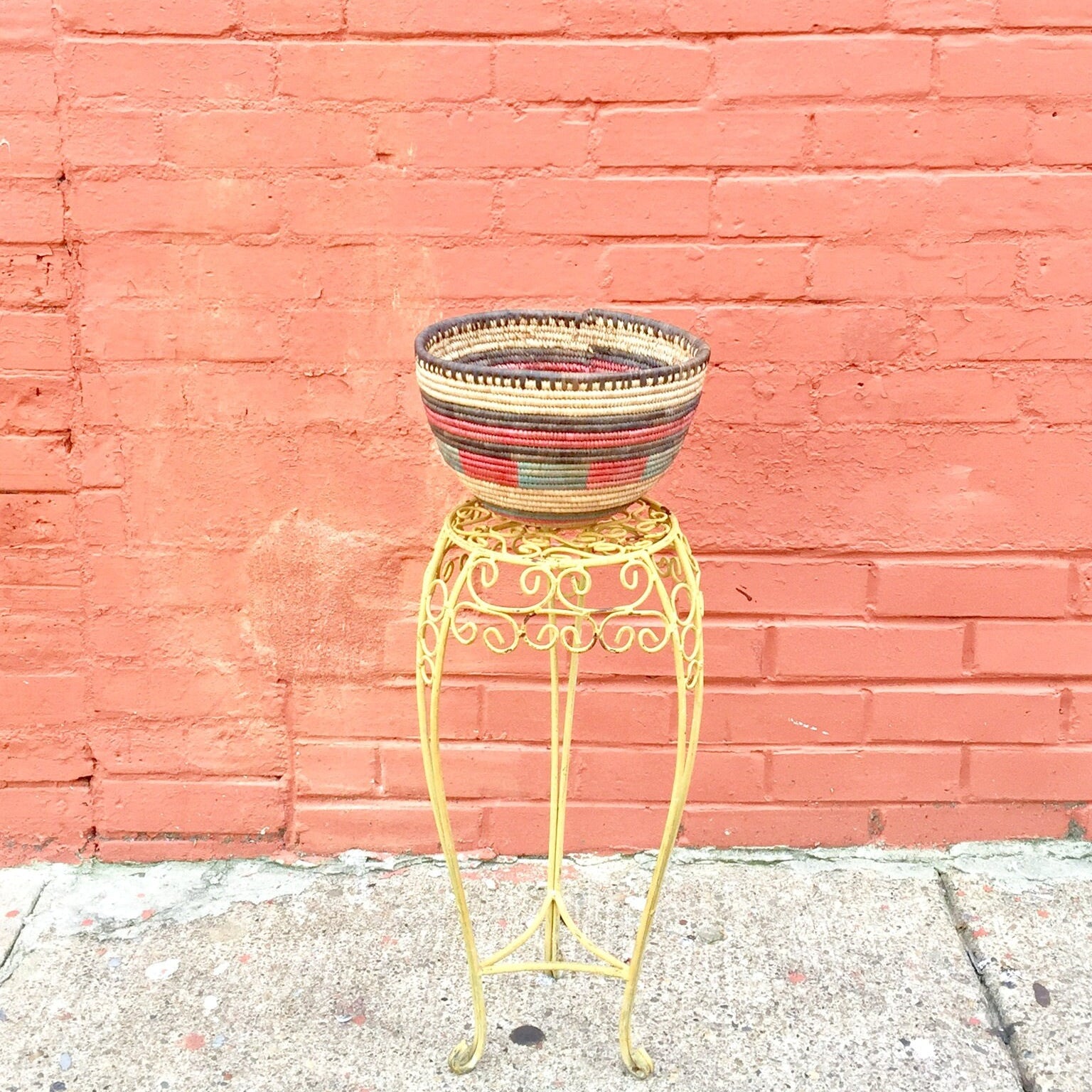 Colorful woven basket on ornate metal stand against pink brick wall, boho vintage home decor