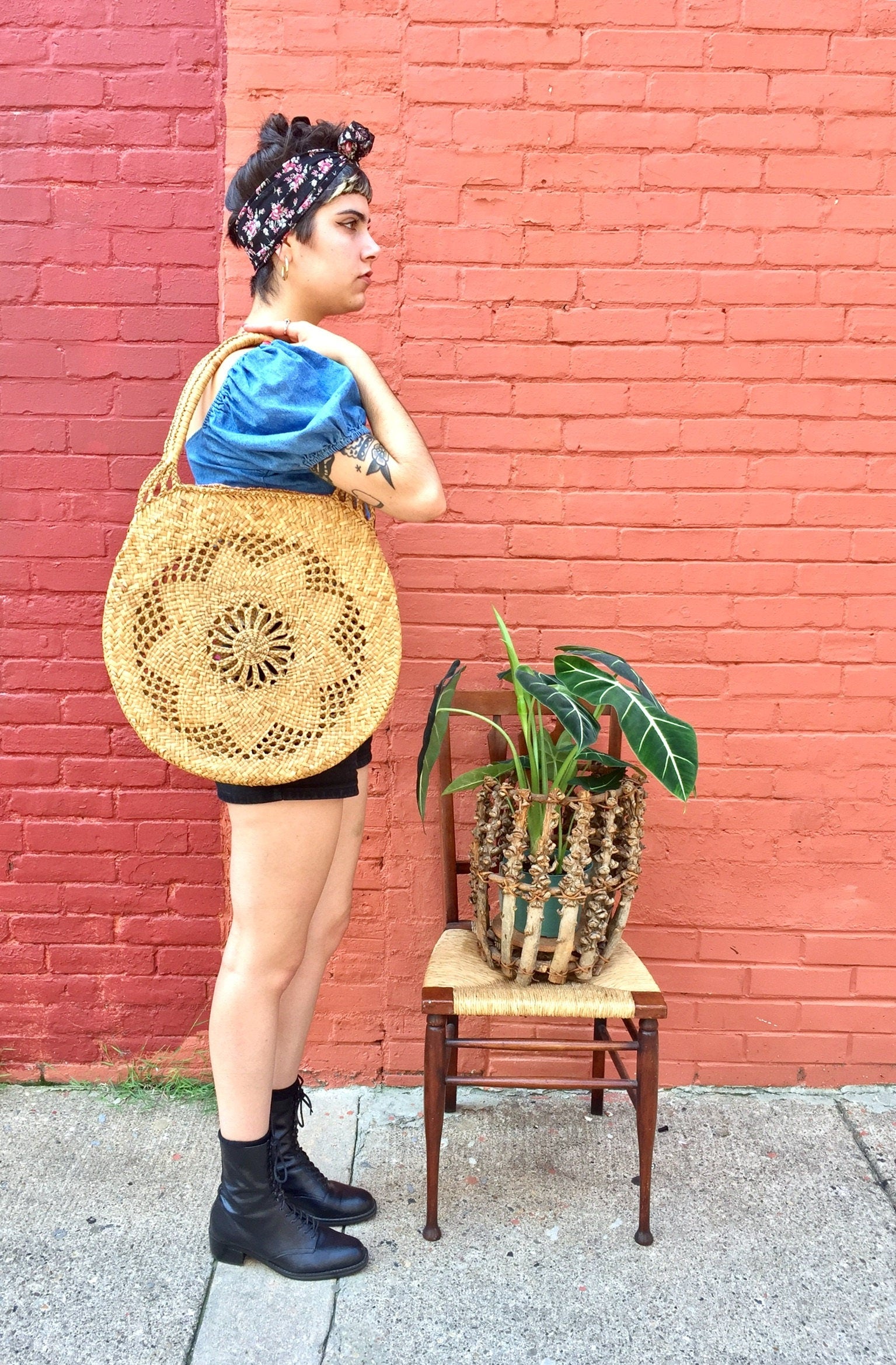 Stylish woman wearing a blue top, patterned yellow tote bag, and black boots standing next to a potted plant in front of a brick wall.