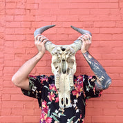 Tattooed person holding a large steer skull adorned with flowers in front of a pink brick wall, showcasing bohemian southwestern home decor.