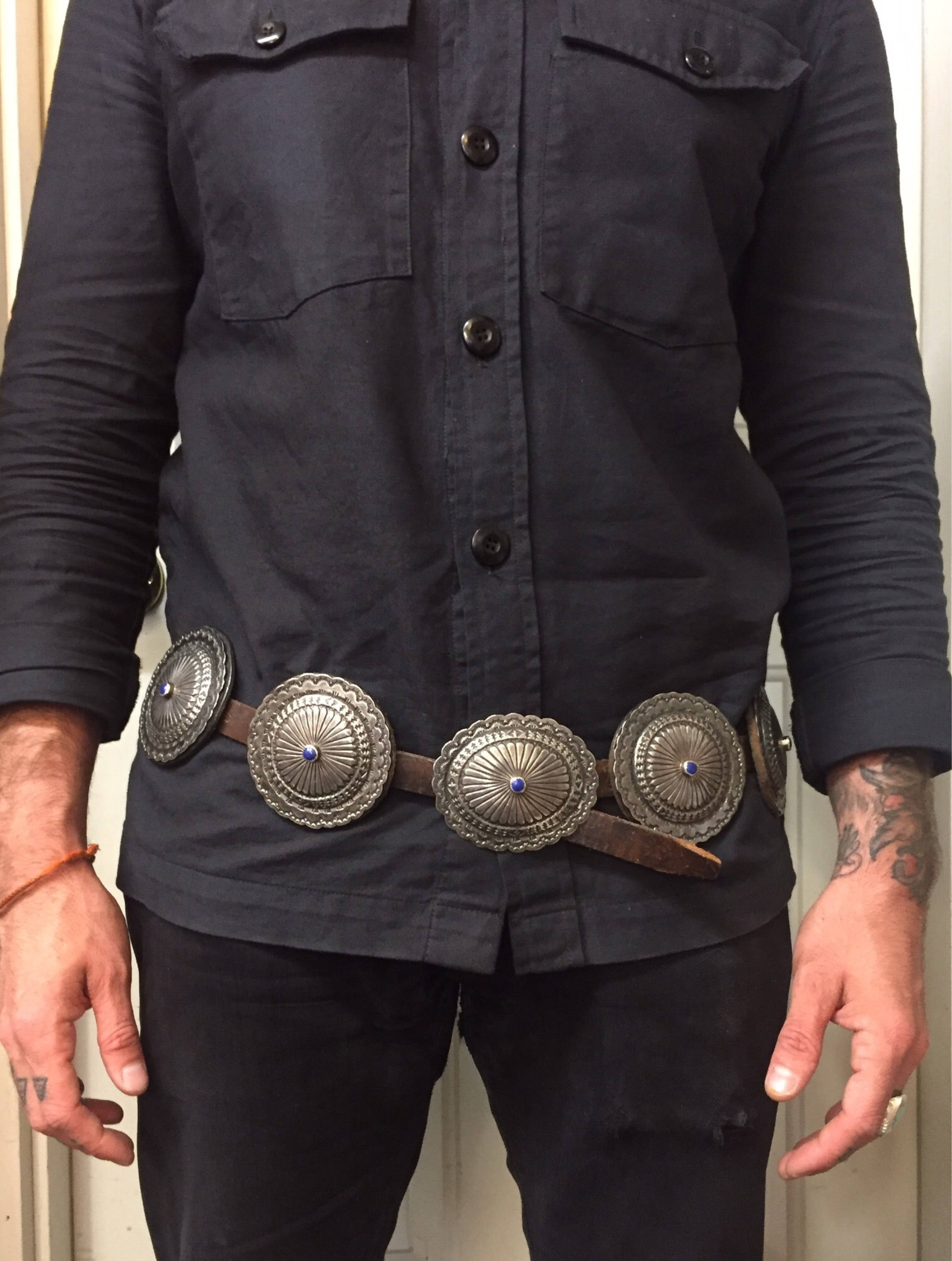 Vintage Navajo handmade sterling silver concho belt with lapis stones, worn by a person in a black button-up shirt and jeans. The large, intricate silver conchos are spaced along a thick leather belt, showcasing traditional Native American silversmithing craftsmanship from the American Southwest.