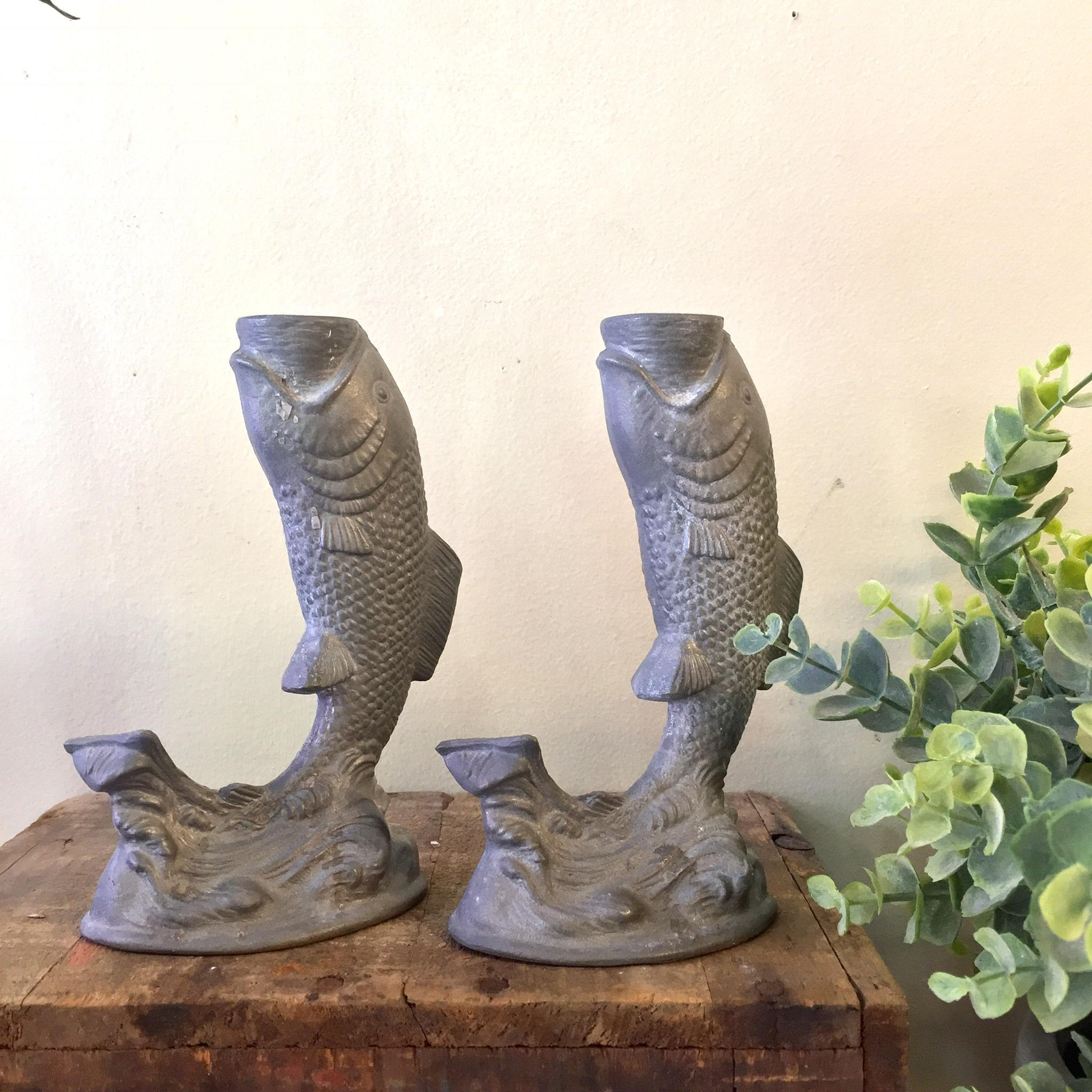 Vintage pewter fish shaped candlestick holders on wooden surface with greenery in background