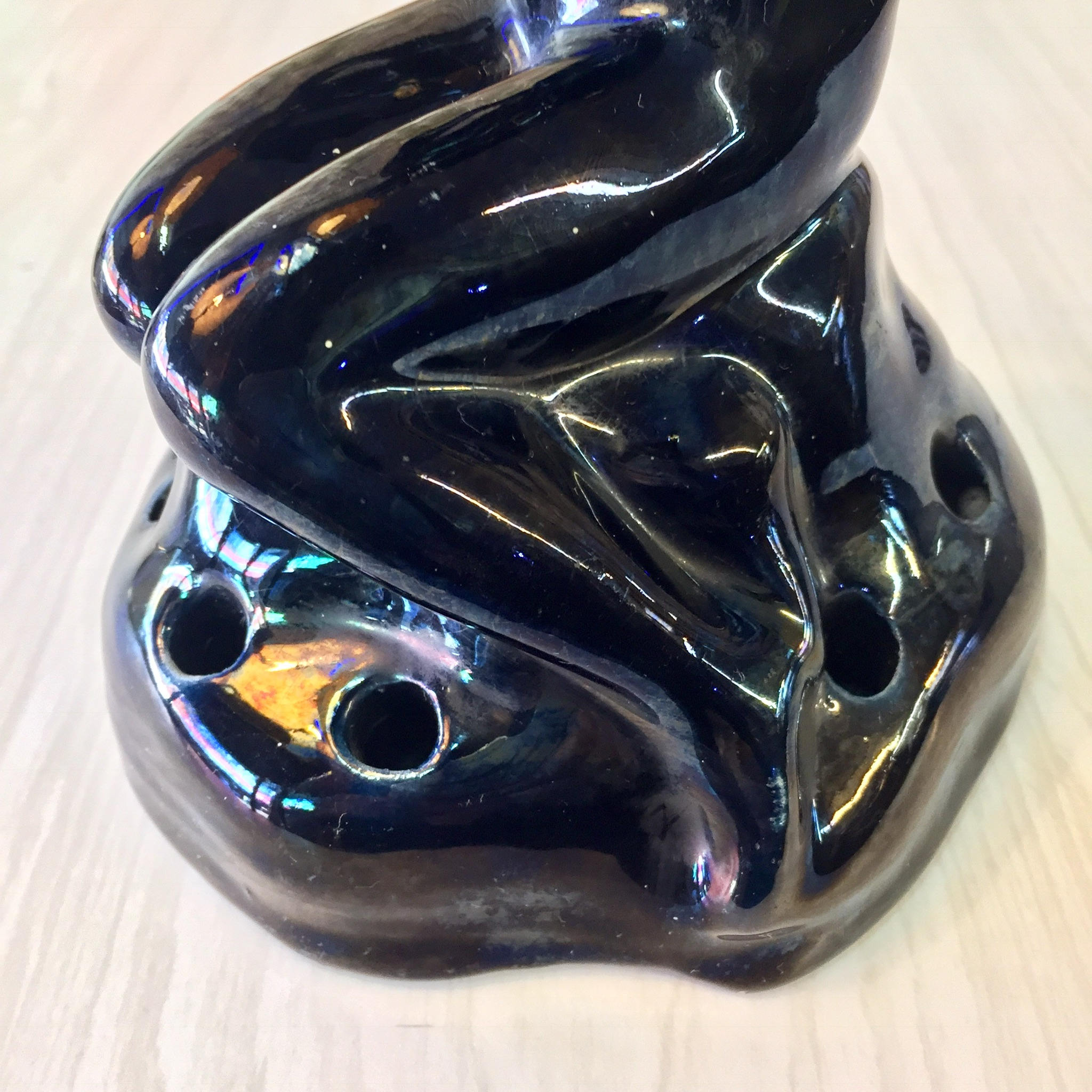 Glossy black ceramic Art Deco style flower frog figurine depicting a stylized nude female form crouching on a rock, with reflective iridescent glaze highlights in blue, green and gold tones.