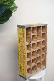 Vintage wooden Coca-Cola bottle crate with 24 compartments, featuring a faded yellow exterior with red Coca-Cola logo and branding text, placed on a white surface next to a green plant leaf.
