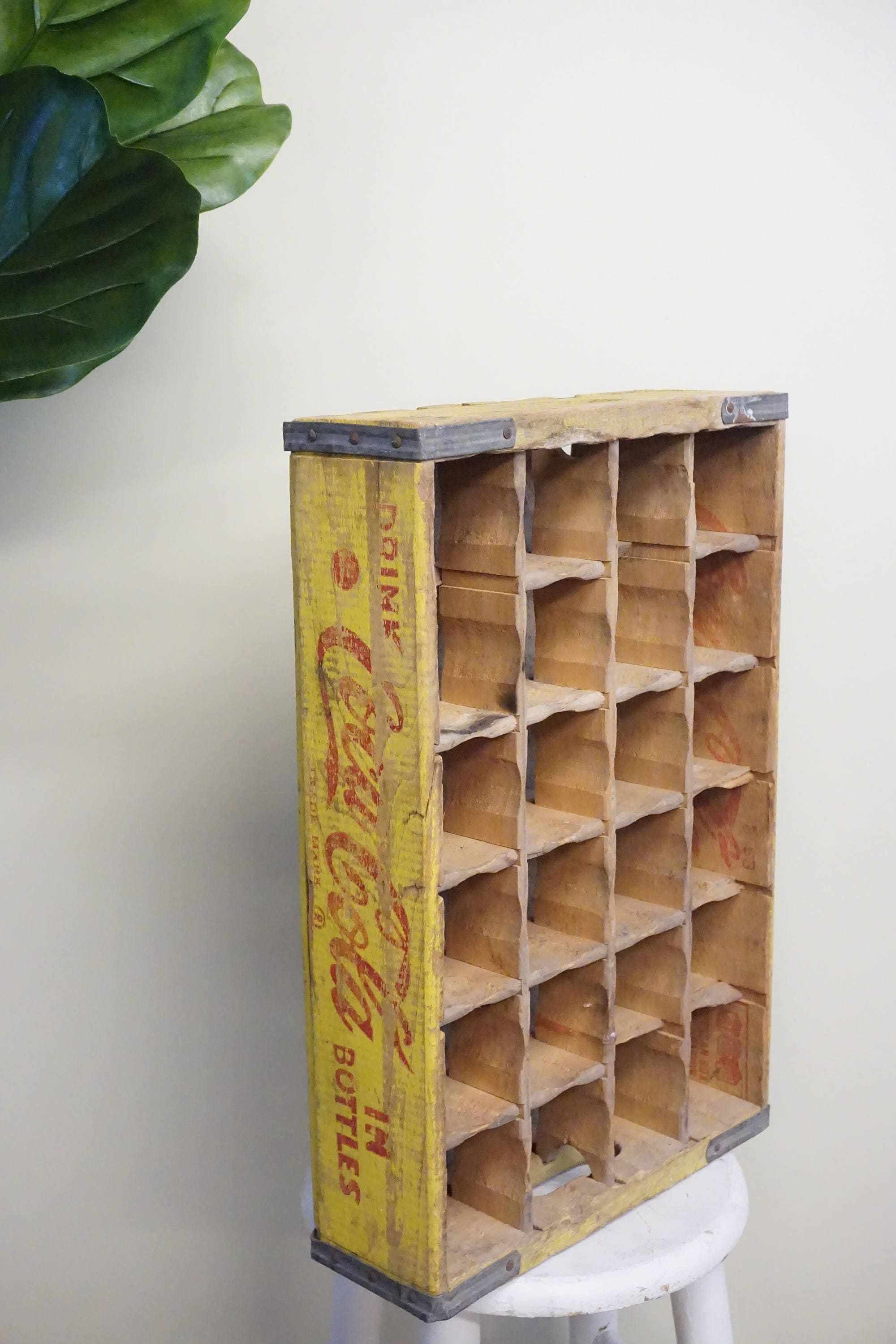 Vintage wooden Coca-Cola bottle crate with 24 compartments, featuring a faded yellow exterior with red Coca-Cola logo and branding text, placed on a white surface next to a green plant leaf.