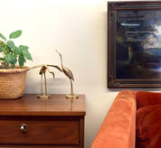 Vintage brass crane statues on wooden table next to orange velvet couch and abstract painting in ornate frame, creating a midcentury modern home decor vignette with brass bird figurines.