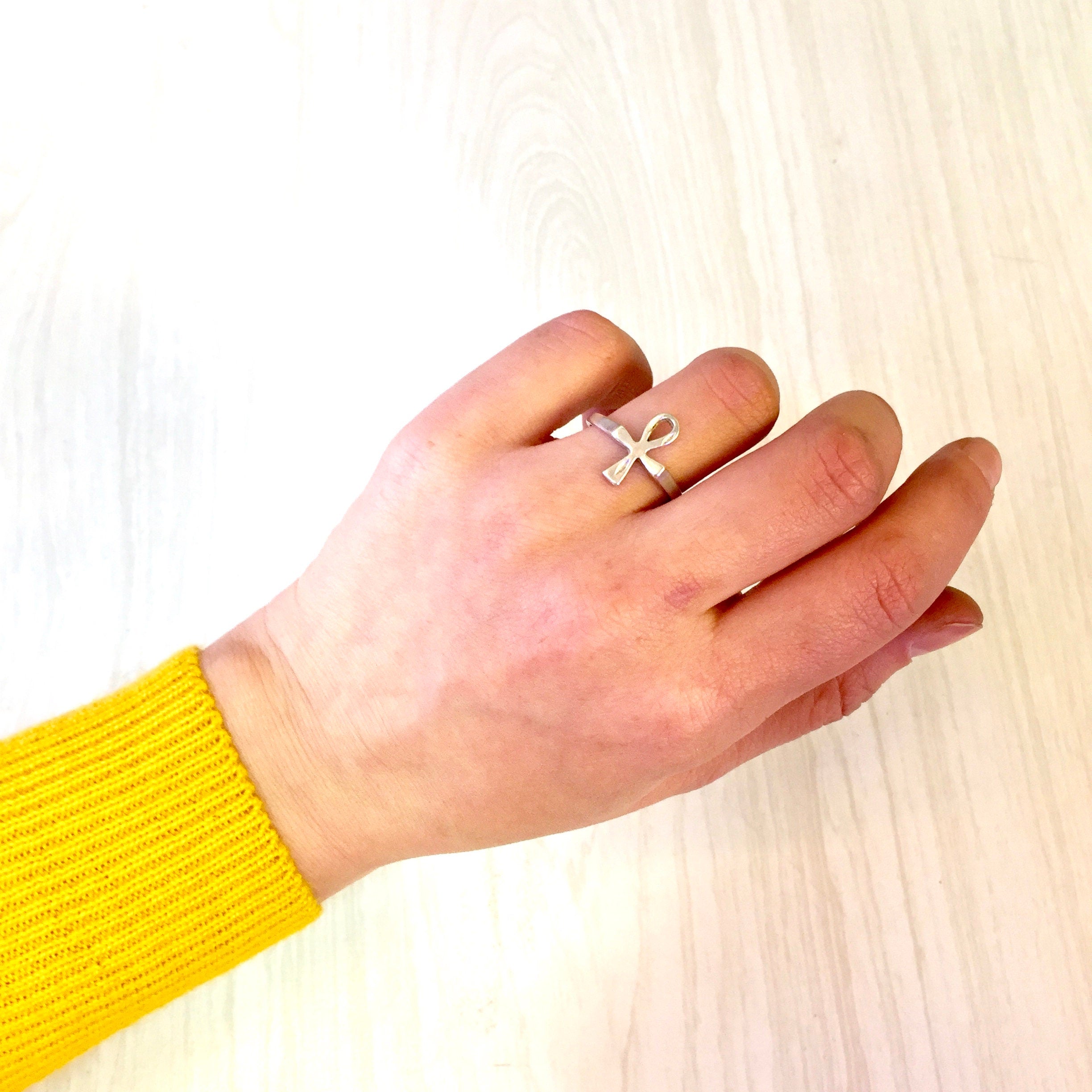 A person's hand wearing a vintage sterling silver ankh ring against a light wood background, with the hand resting on a bright yellow sweater sleeve.