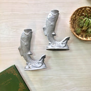 Vintage pewter fish-shaped candlestick holders on wooden surface next to woven basket and green book, suitable for holiday gift or home decor.