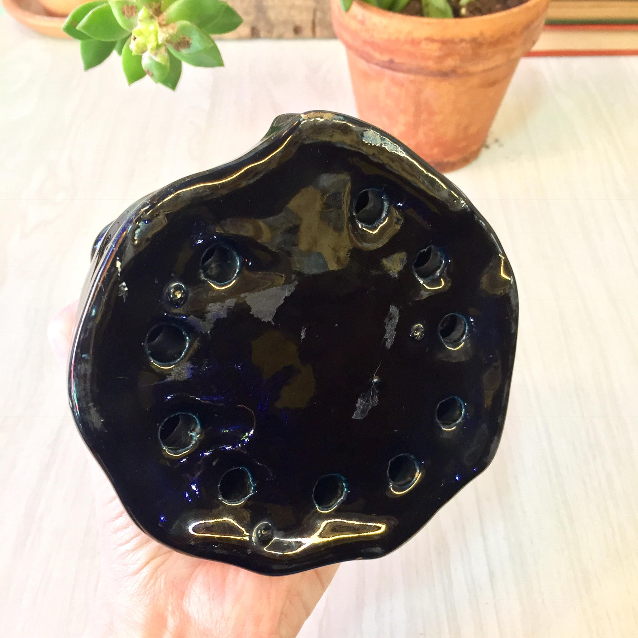 Vintage black ceramic flower frog shaped like an abstract nude woman, with glossy glaze and bubble textures, held in a hand in front of potted plants on a white wooden surface.