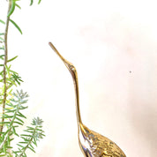 Brass crane statue with elongated neck and beak, perched amidst green fern foliage, against a plain white background. Vintage midcentury modern brass bird figurine for home decor.
