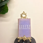 Vintage brass easel book holder with intricate filigree design, displayed with a purple book titled "Vogue's Book of Etiquette", on a white background with green foliage.