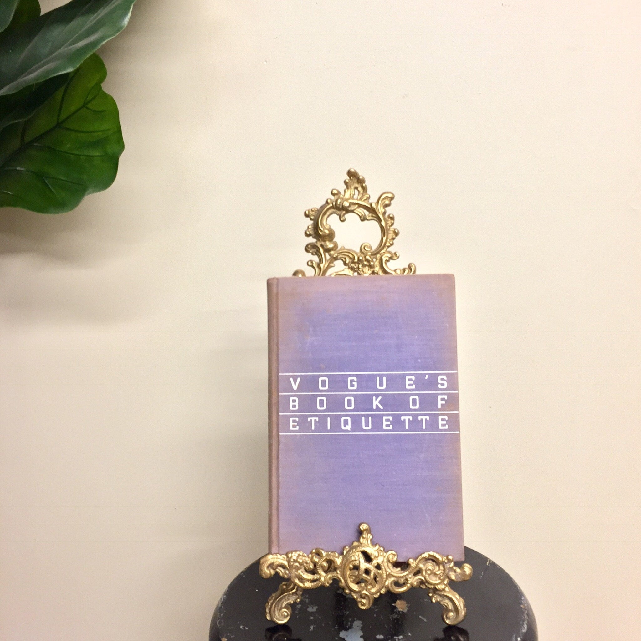 Vintage brass easel book holder with intricate filigree design, displayed with a purple book titled "Vogue's Book of Etiquette", on a white background with green foliage.