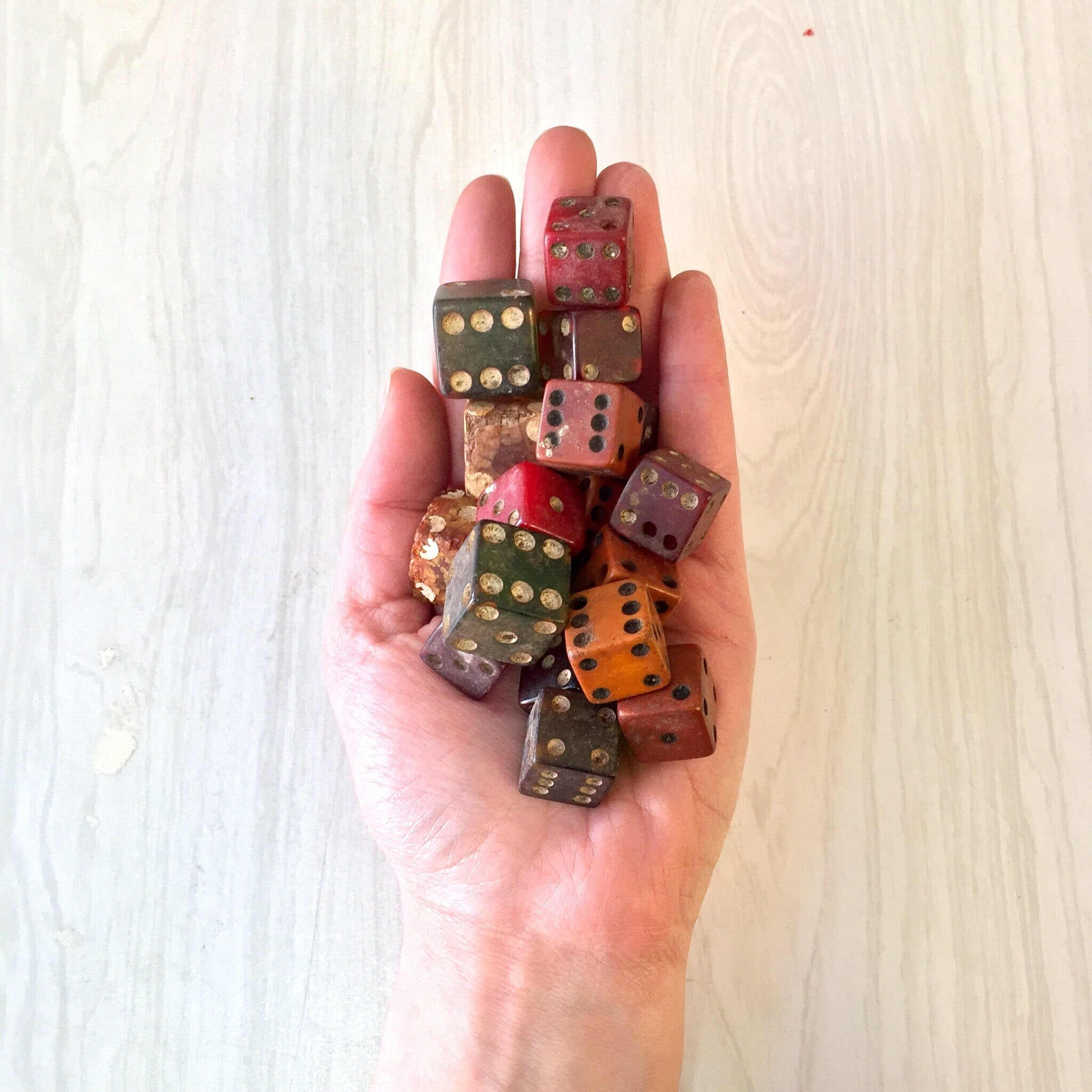 A hand holding a large collection of antique dice in various colors and styles, including wooden, metal and plastic dice, some showing signs of wear from use in games or crafting projects.
