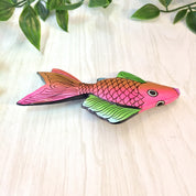 Large wooden fish-shaped brooch pin with pink, orange, green, and blue colors against a light wooden background with green leaves.