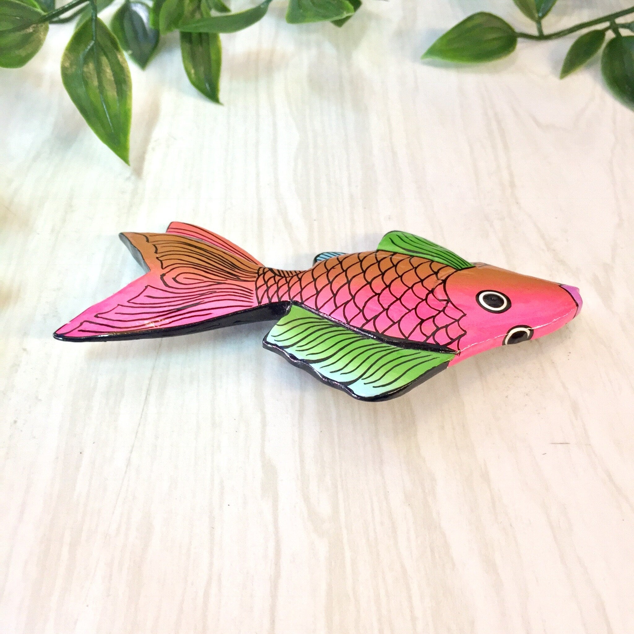Large wooden fish-shaped brooch pin with pink, orange, green, and blue colors against a light wooden background with green leaves.