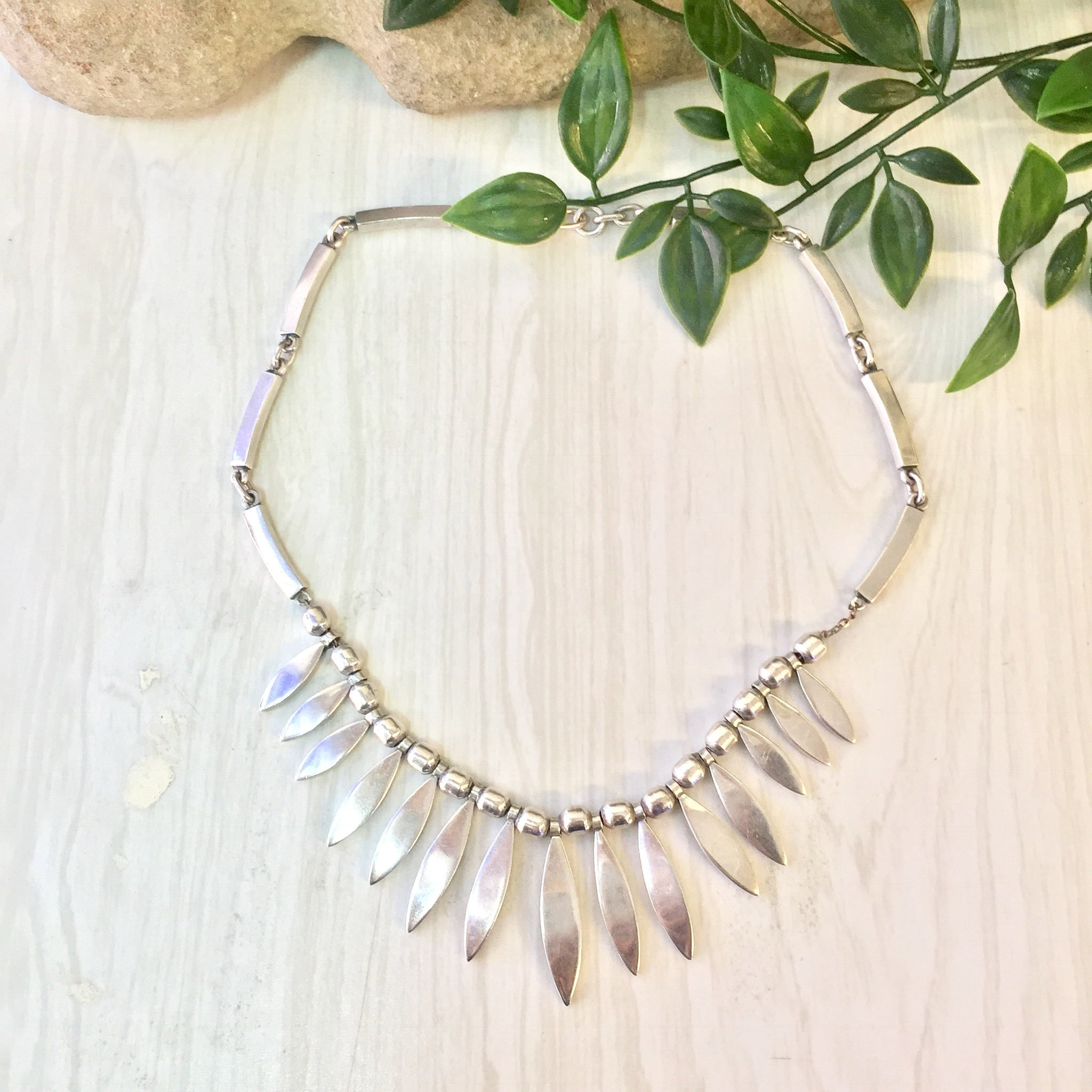 Vintage sterling silver statement bib necklace with dangling leaf pendants, displayed on a wooden surface with green foliage in the background.