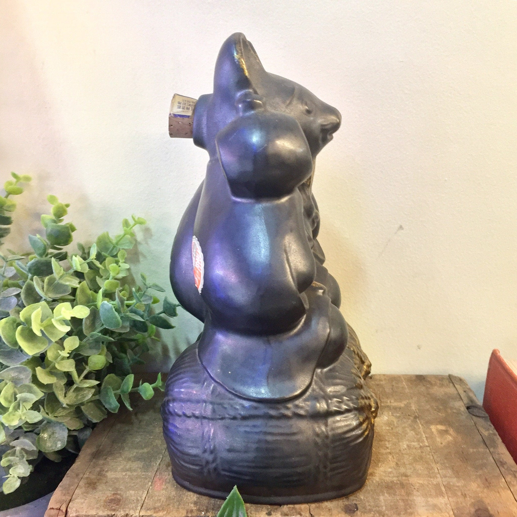 Vintage black and gold ceramic sake bottle shaped like a seated rat or mouse figure on a wooden surface with greenery in the background.