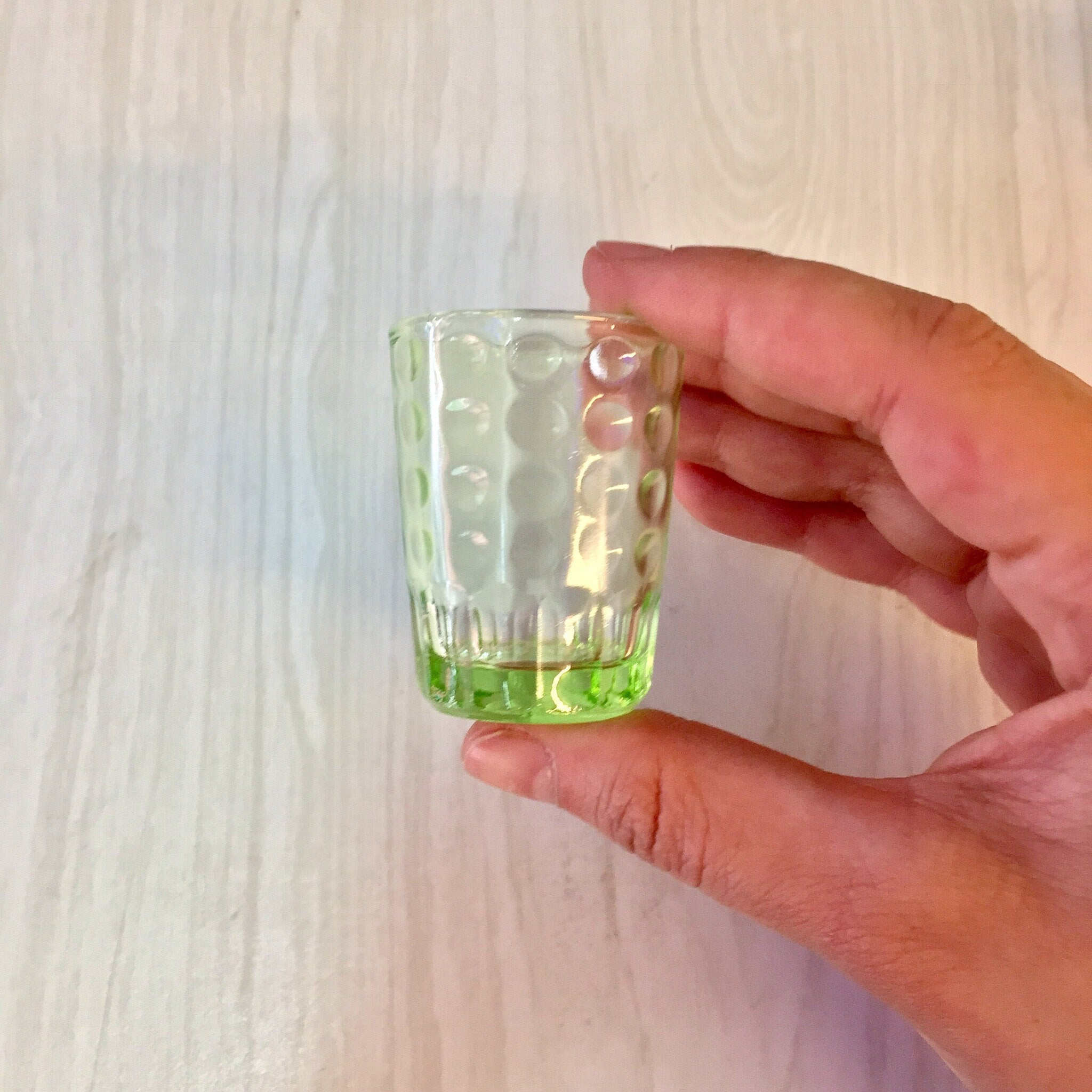 Hand holding a small, transparent green glass shot glass with a polka dot pattern against a white wooden background.