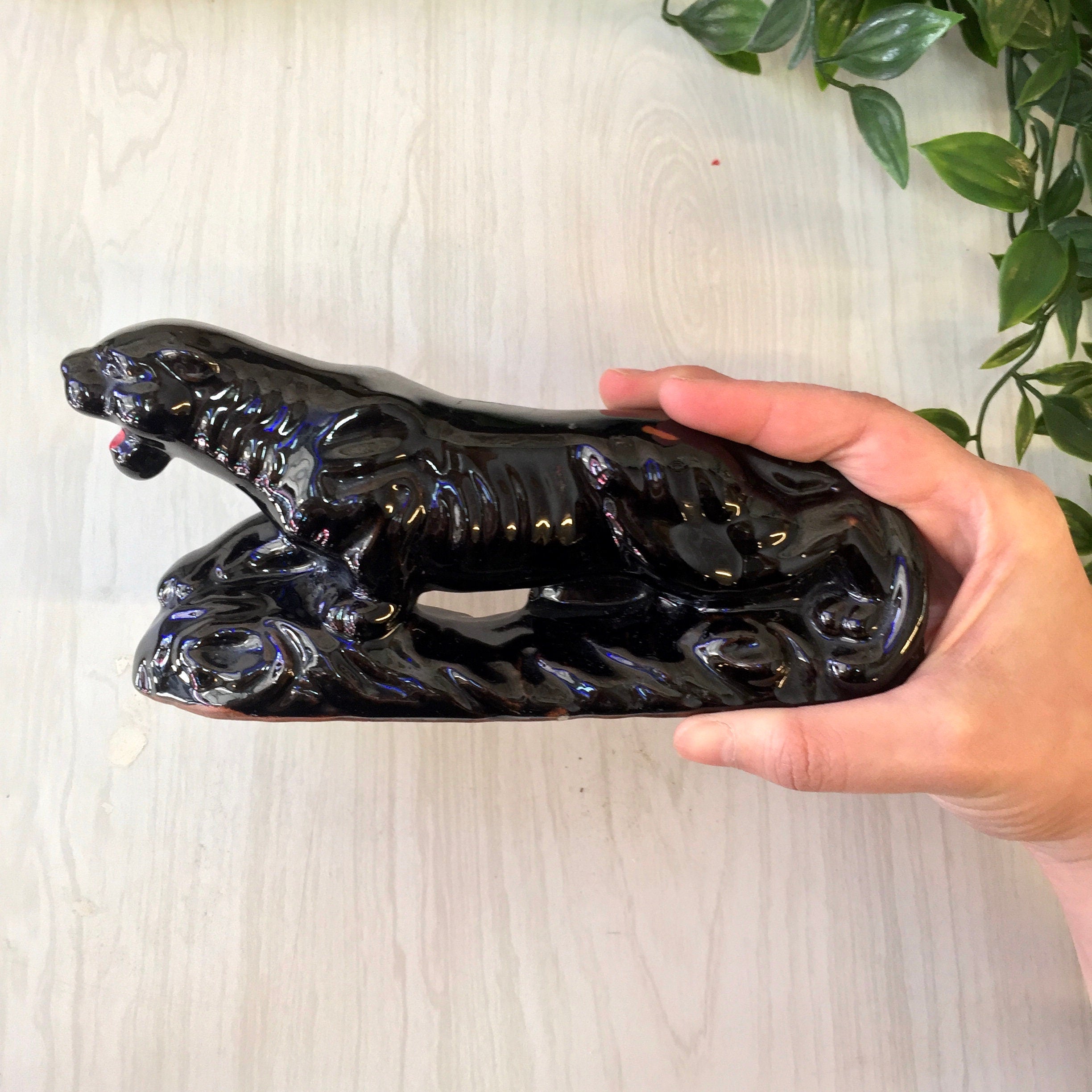 Hand holding a vintage black ceramic panther figurine with glossy glaze, against a light wooden background with a plant visible on the right side of the frame.