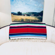 Vintage Mexican blanket with vibrant stripes of red, blue, green and pink draped over a chair, with a fringe tassel edge and wheat field landscape painting in the background.