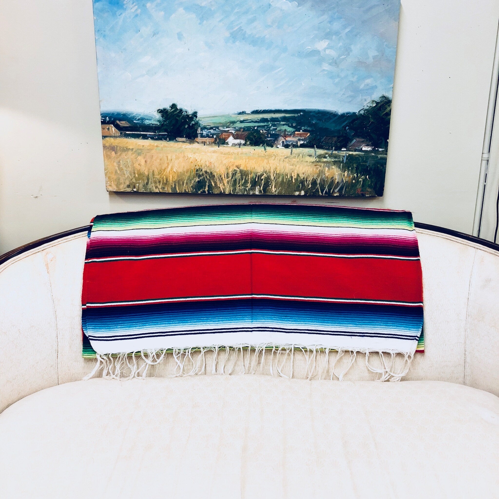 Vintage Mexican blanket with vibrant stripes of red, blue, green and pink draped over a chair, with a fringe tassel edge and wheat field landscape painting in the background.