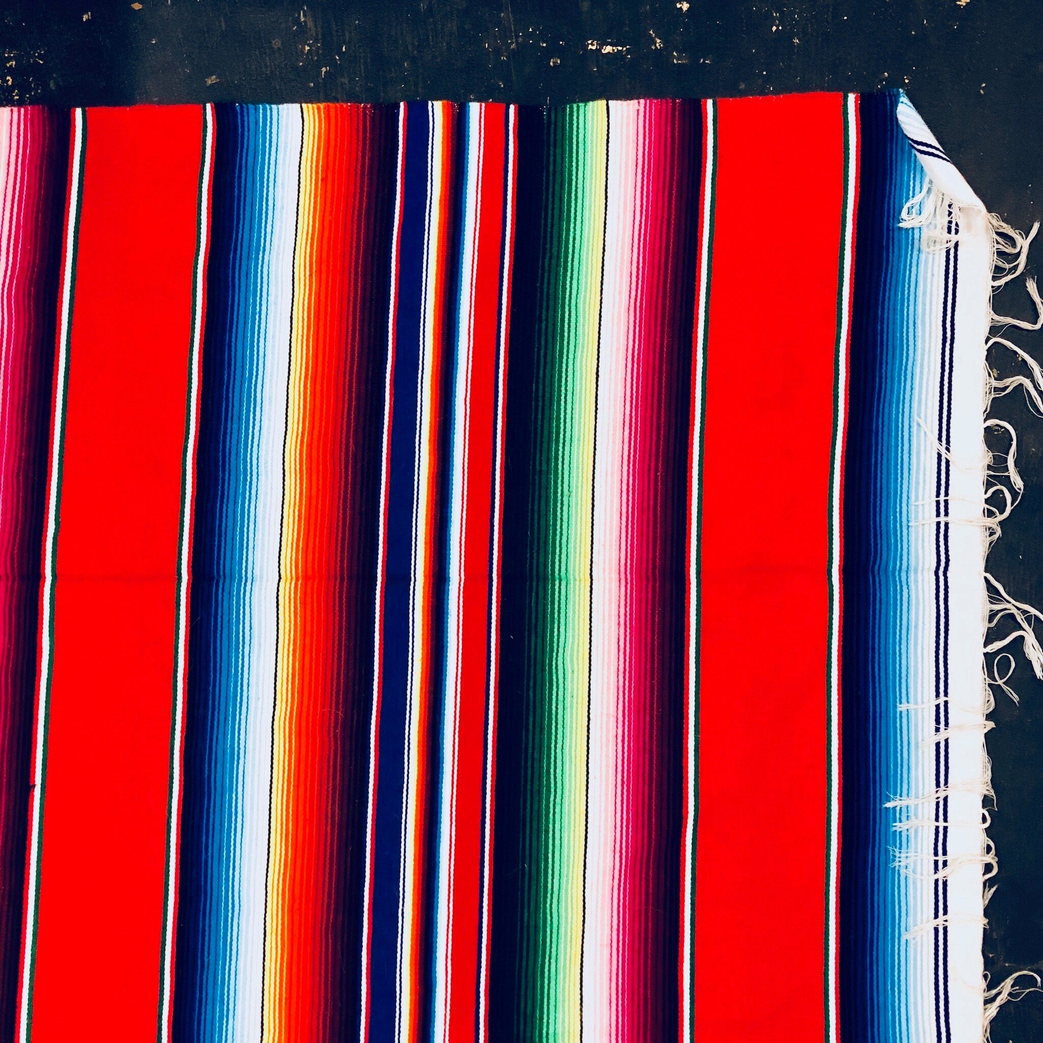 Colorful vintage Mexican blanket with red, blue, green, pink and white stripes, featuring tasseled fringes, suitable for use as a throw or picnic blanket.