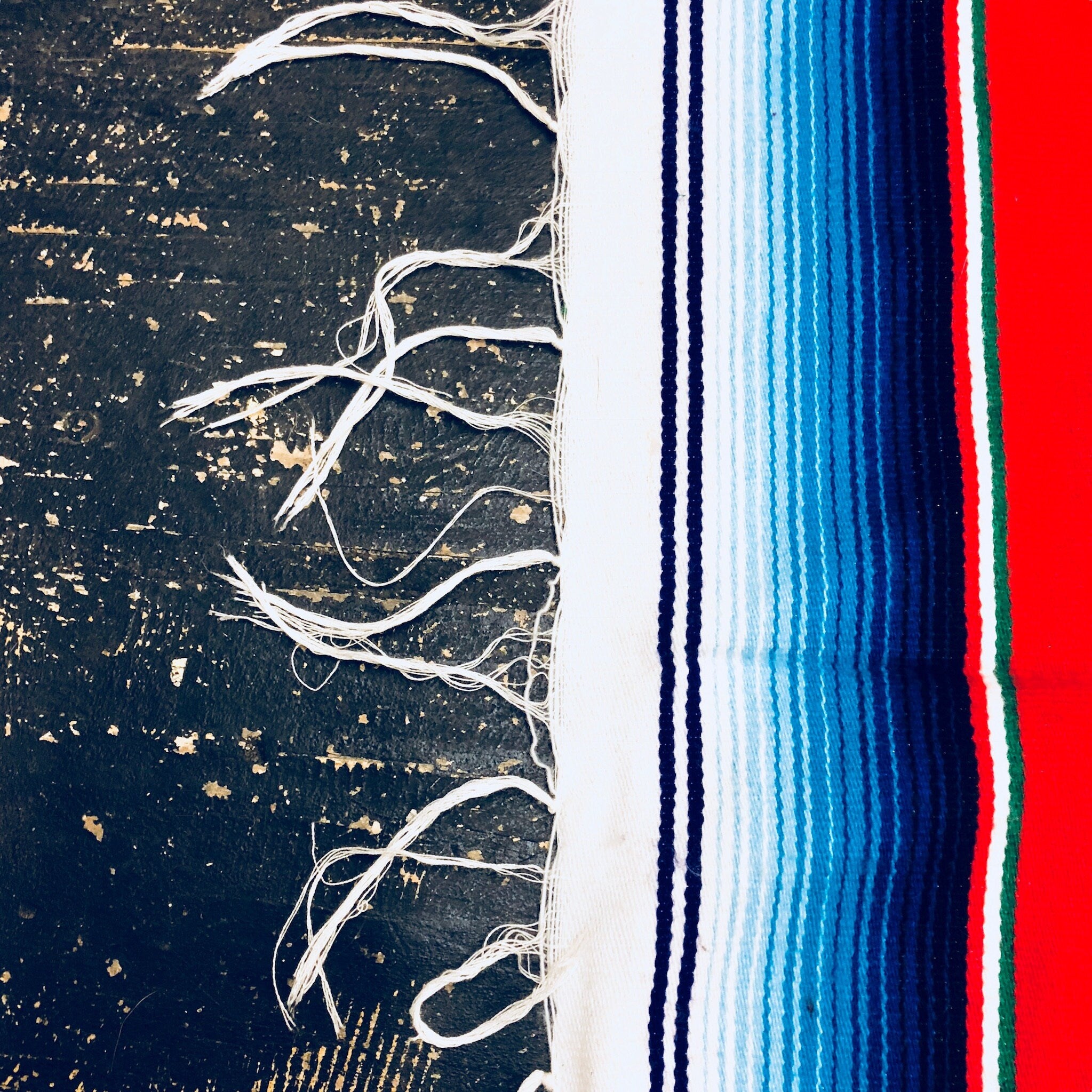 Vintage Mexican blanket with bright stripes of red, blue, green and white on dark background, fringed edges, close-up detail of textured woven fibers and tassels