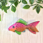 Colorful vintage wooden fish brooch pin with pink, orange, and green scales against a wooden background with green leaves