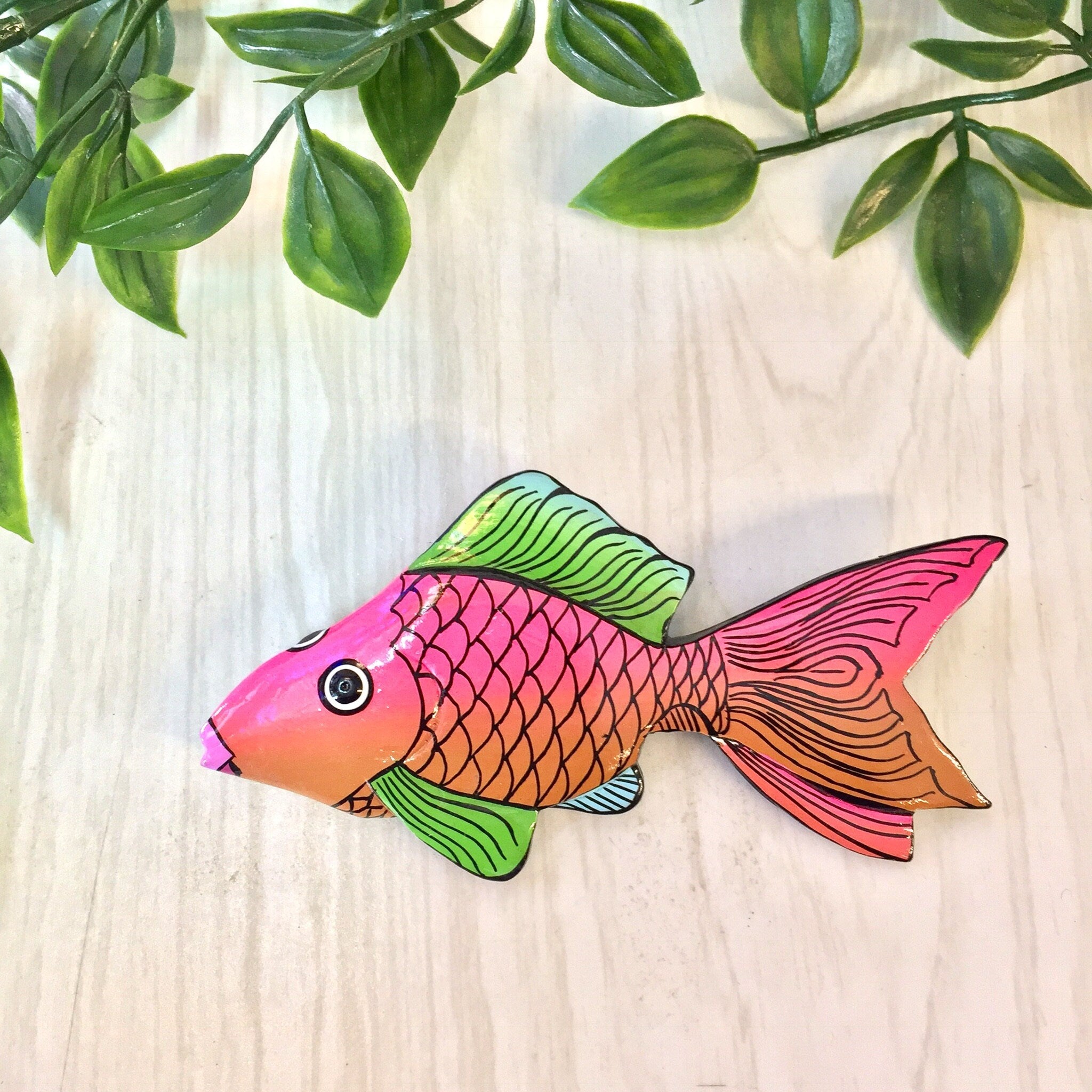 Colorful vintage wooden fish brooch pin with pink, orange, and green scales against a wooden background with green leaves