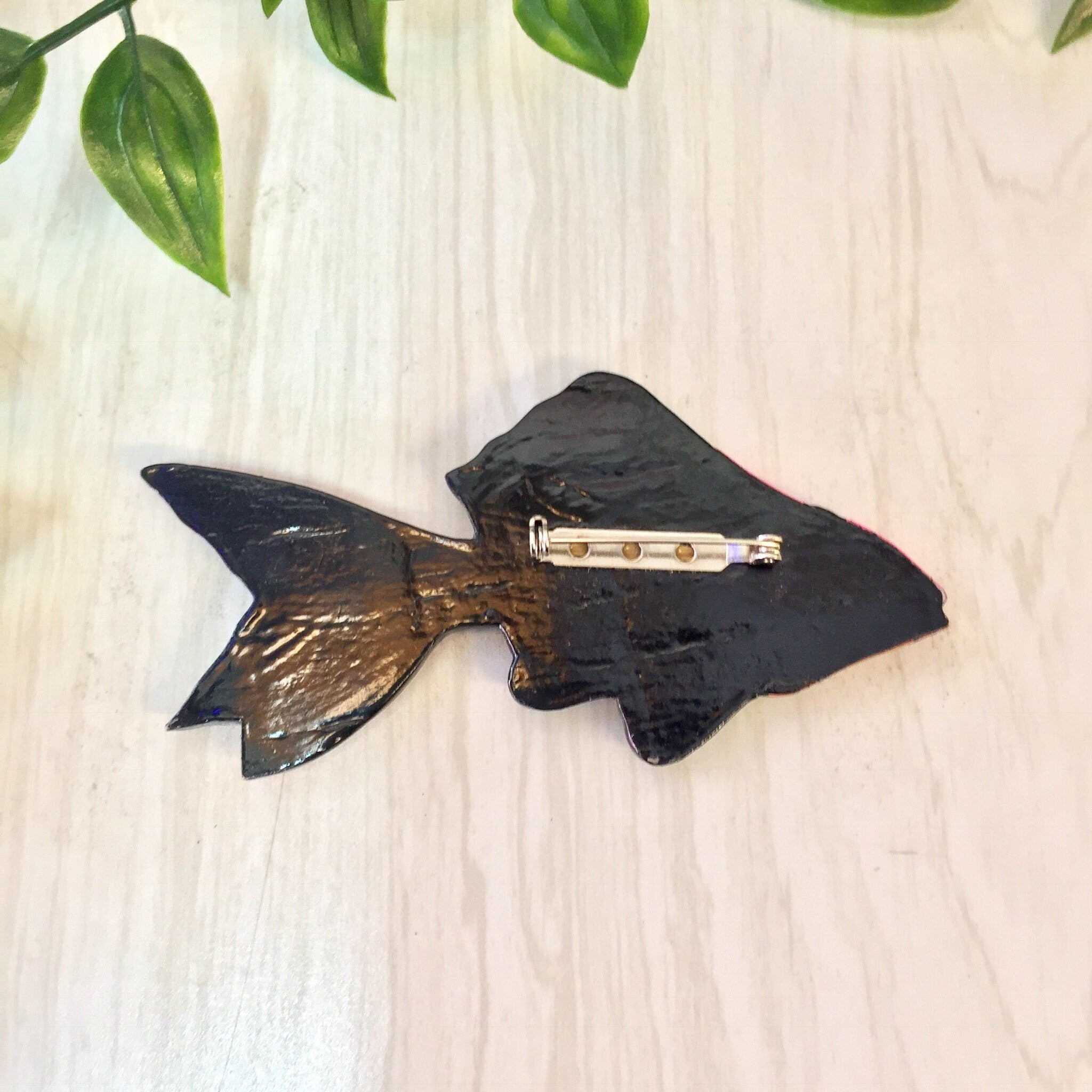 Large wooden fish brooch pin with pink, orange, green and blue colors on wooden background with green leaves