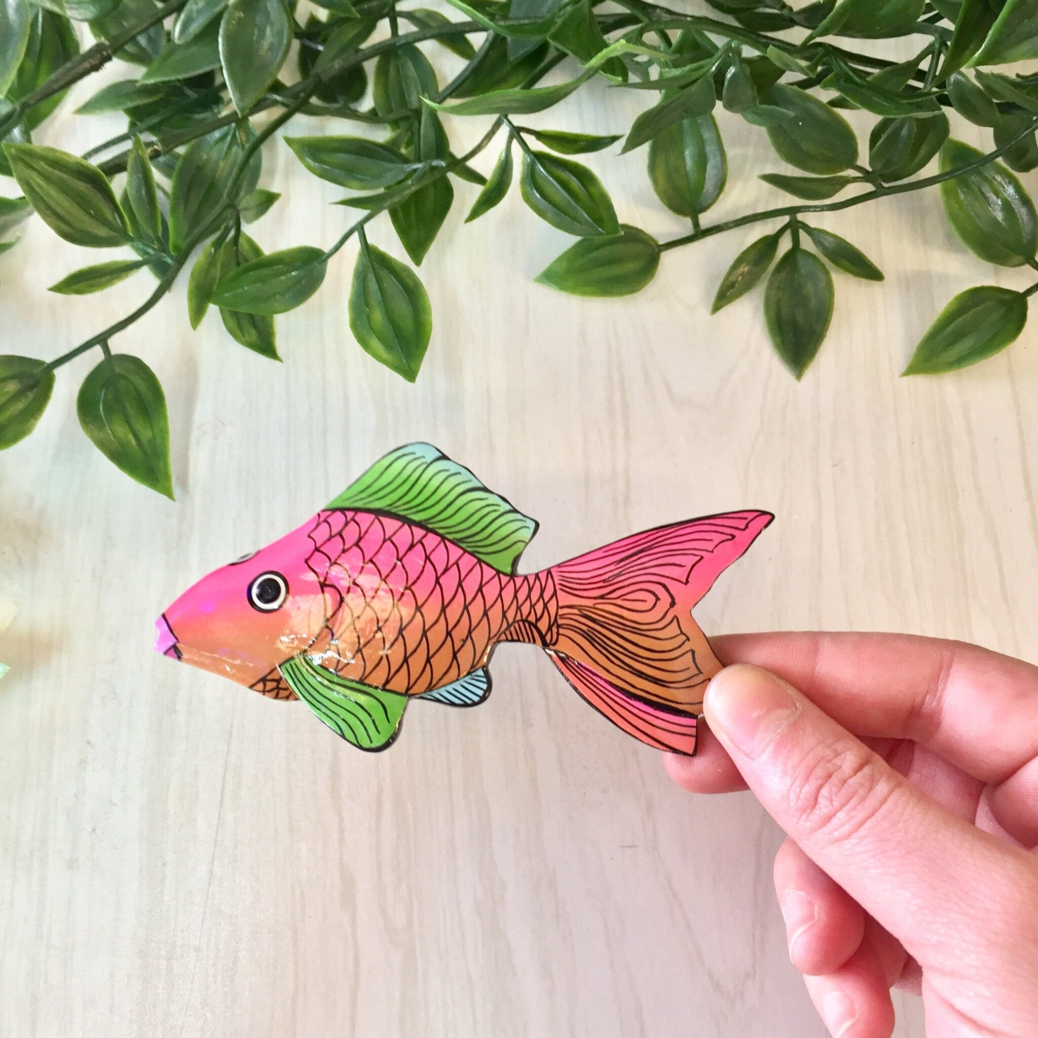 Hand holding a colorful wooden fish brooch pin featuring pink, orange and green colors against a background of green leaves.