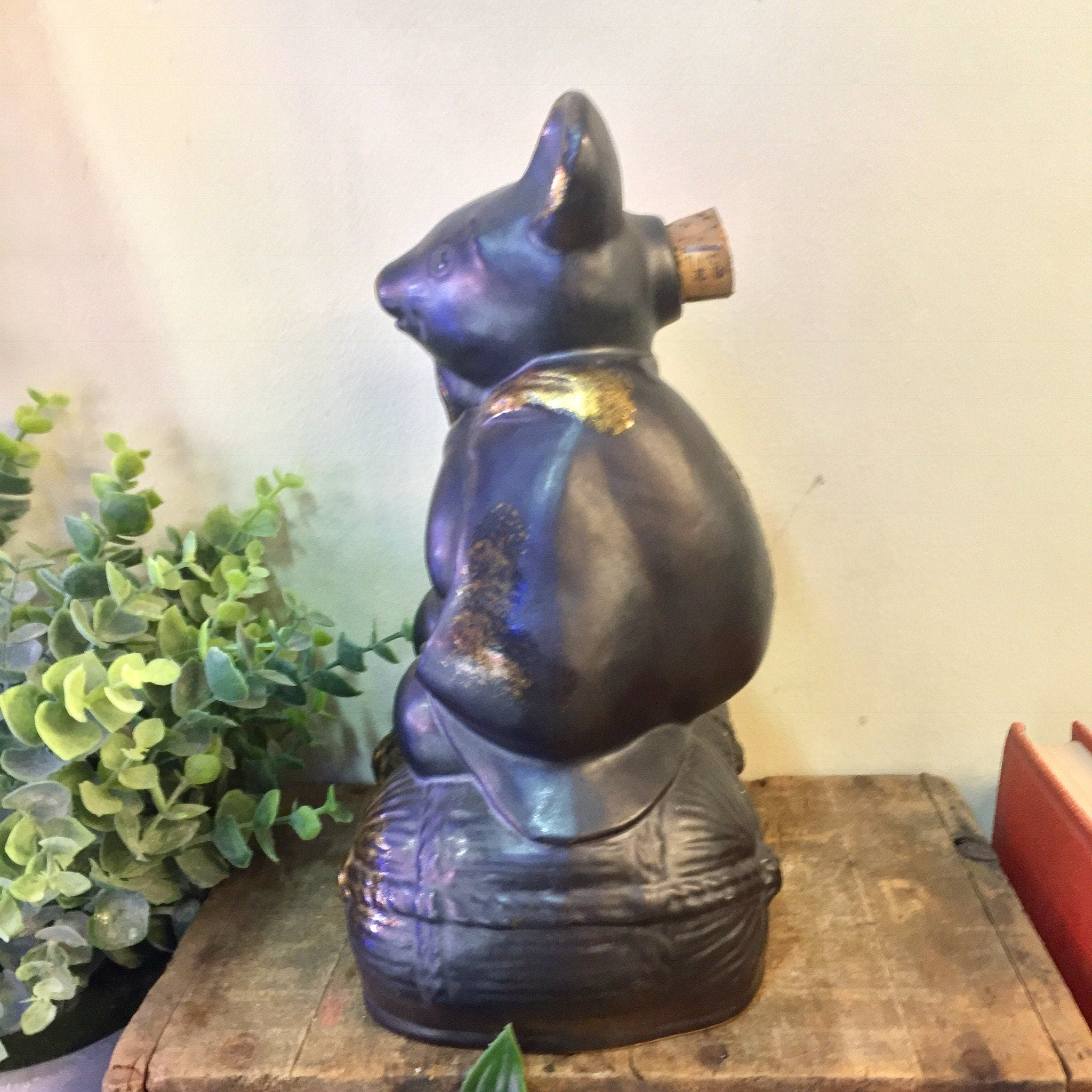 Vintage blue and gold glazed ceramic sake bottle in the shape of a sitting rat or mouse figurine, displayed on a wooden surface with greenery in the background.