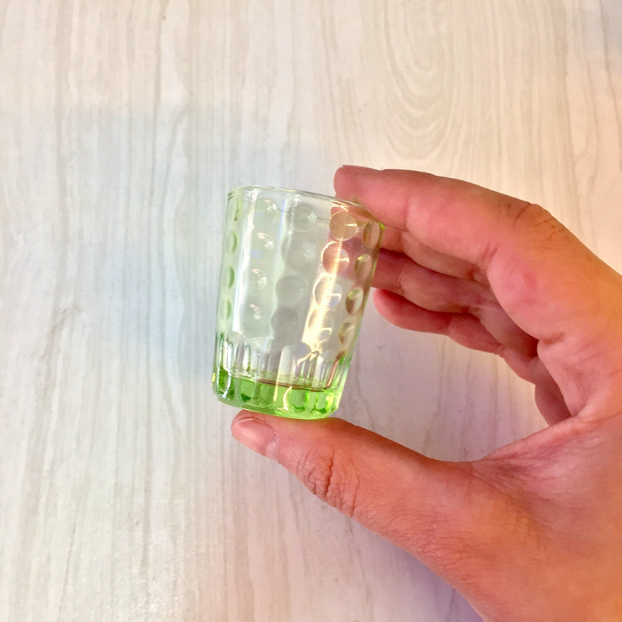 Hand holding a green vintage polka dot depression glass shot glass against a wooden background