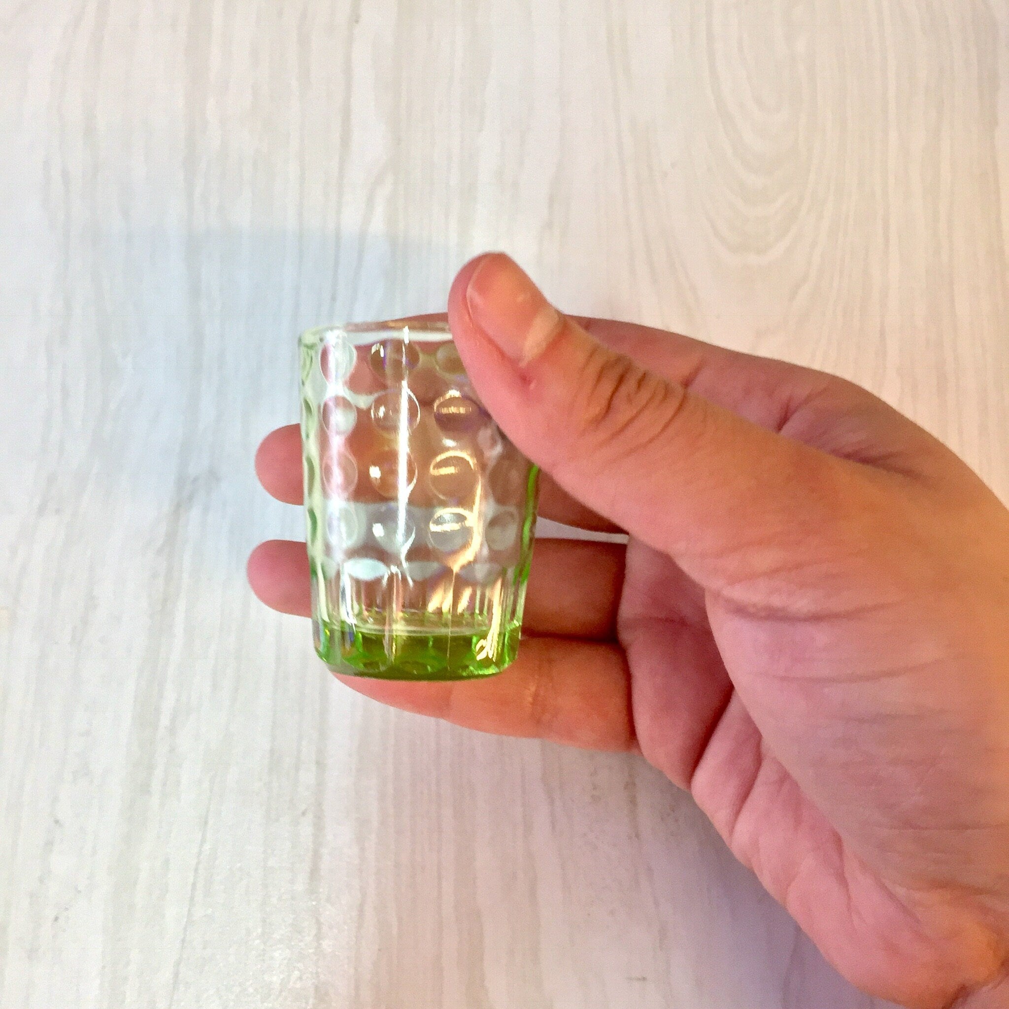 Hand holding a green vintage depression glass shot glass with a polka dot design against a light wooden background.