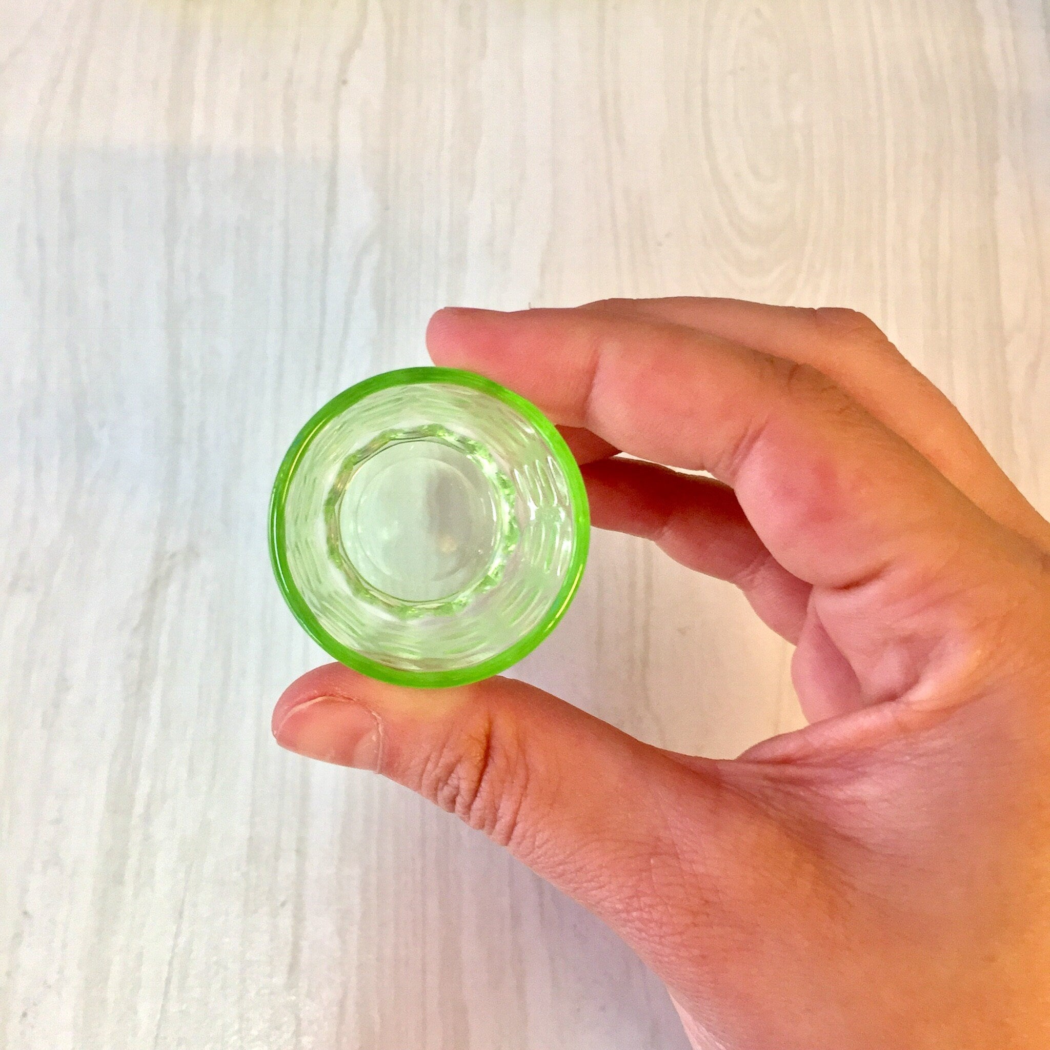 Hand holding a small, translucent green glass shot glass with a polka dot design against a light wood background.
