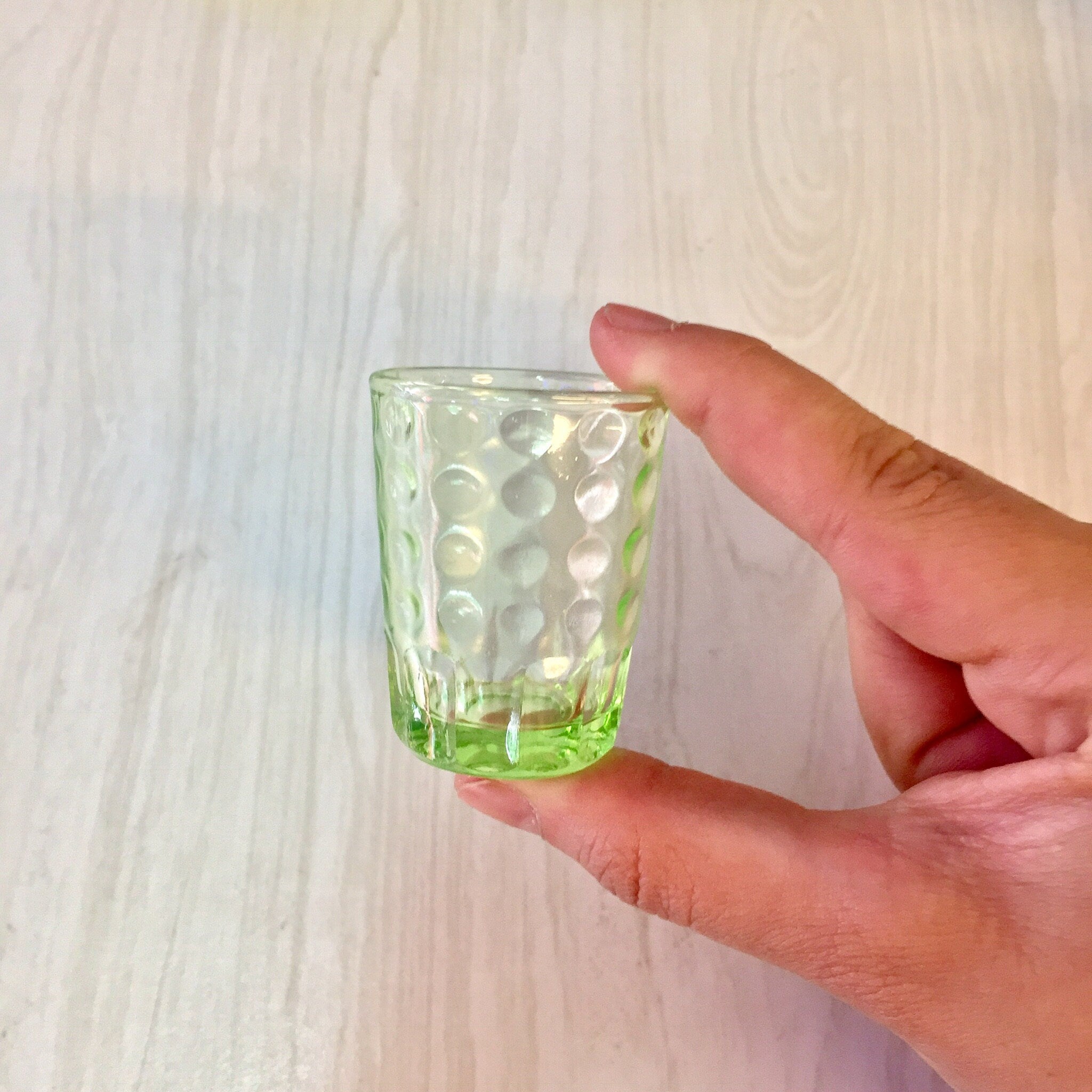 Hand holding a small, vintage green depression glass shot glass with a polka dot design against a light wood background.