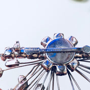 Close-up of a vintage Marvella jewelry set featuring a silver brooch with blue and clear rhinestones and matching clip-on earrings, showcasing an art deco design.