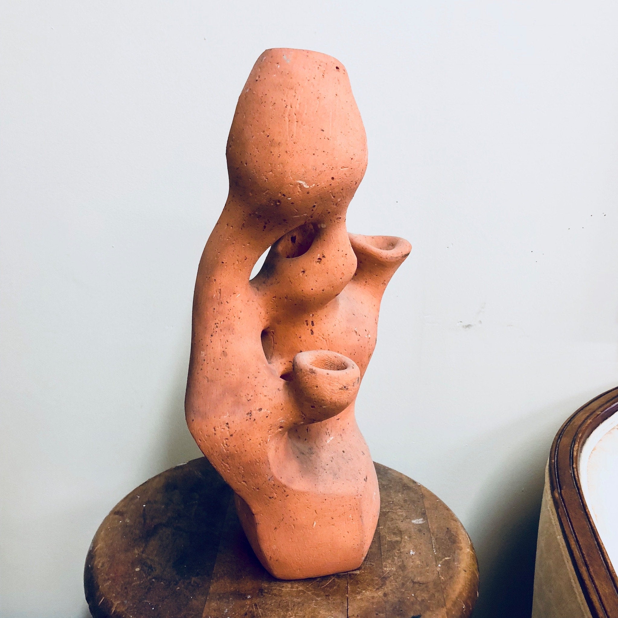 Terracotta abstract sculpture on wooden stand depicting two interlocking human-like figures in an intimate embrace, with pitted and textured surface suggesting aging or weathering.