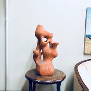 Terracotta sculpture of two abstract human figures embracing on a wooden stool, with paintings hanging on the wall in the background.