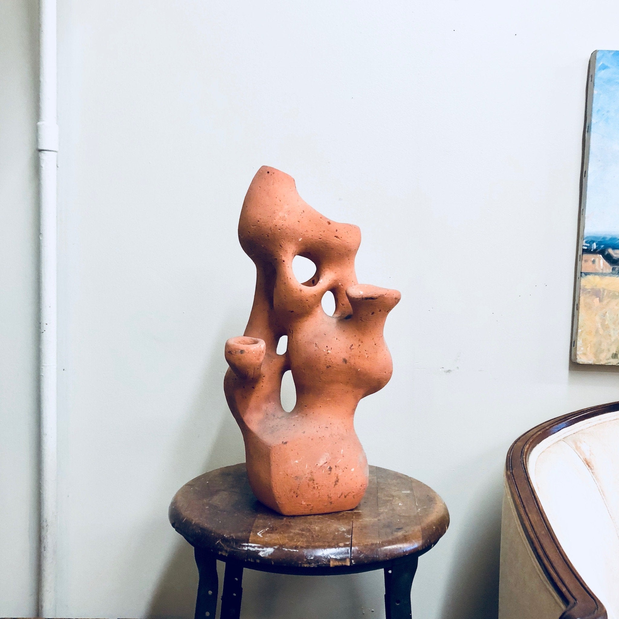 Terracotta sculpture of two abstract human figures embracing on a wooden stool, with paintings hanging on the wall in the background.