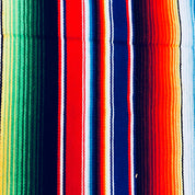 Colorful vintage Mexican blanket with vertical stripes in vibrant shades of red, blue, green, pink, and white, featuring fringed edges and tassels at the ends, suitable for use as a throw or picnic blanket.