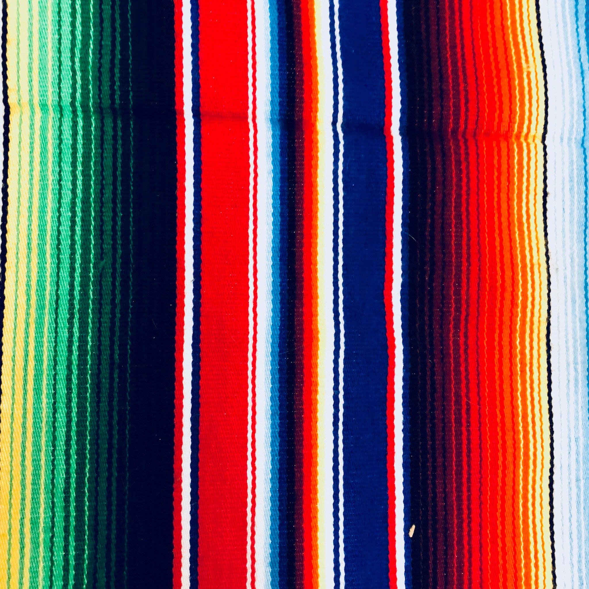Colorful vintage Mexican blanket with vertical stripes in vibrant shades of red, blue, green, pink, and white, featuring fringed edges and tassels at the ends, suitable for use as a throw or picnic blanket.