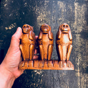 Wooden carved figurines of the three wise monkeys sitting on a hand, representing "see no evil, hear no evil, speak no evil", against a distressed dark background.