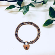 Vintage gold-toned chain bracelet with heart-shaped locket charm, displayed on white fabric with green leaf accents in the background.