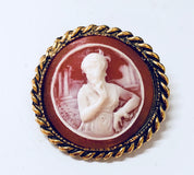 Vintage gold-toned cameo brooch featuring a white carved figure against a dark orange background, surrounded by a twisted rope border.